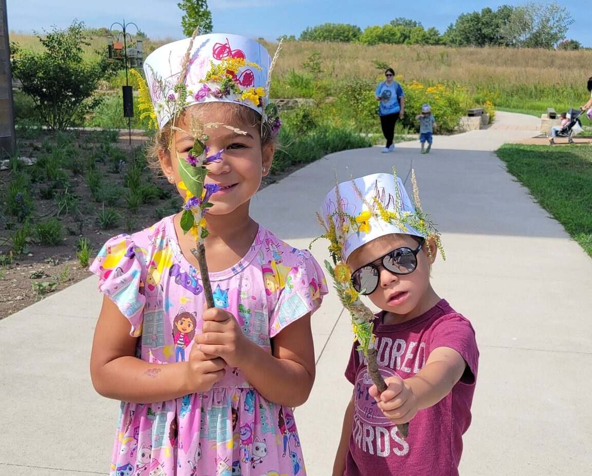 Two children holding stick wands and wearing crowns made of natural materials.