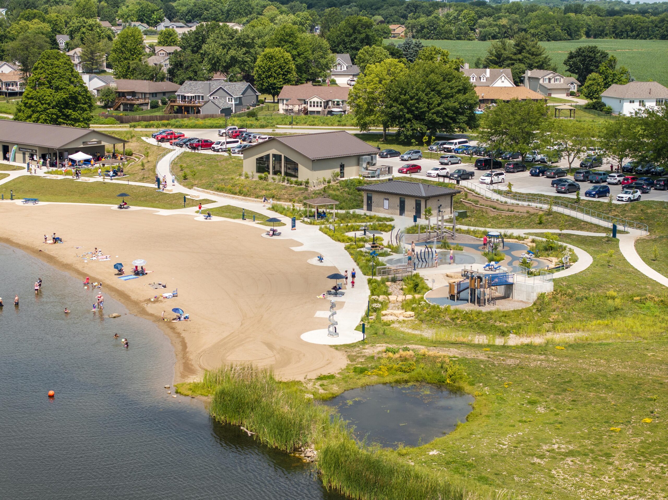 Arial shot of Athene North Shore Recreation Area. This image include the Events building, concessions, rentals, playground, beach, and parking lot. There are people scattered about recreating.