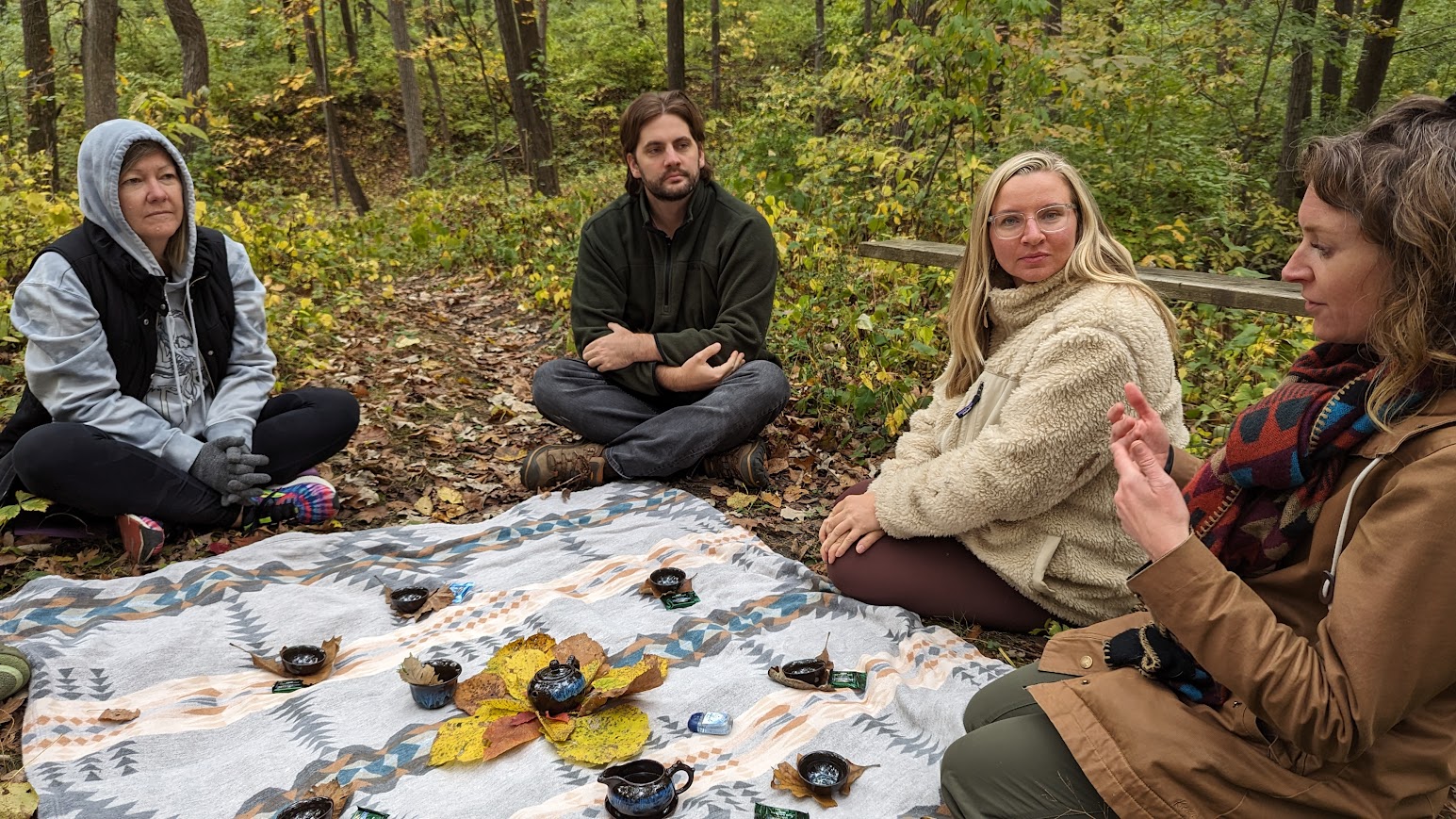 Image of a group of four people sitting on the ground in a forest with a blanket under them.